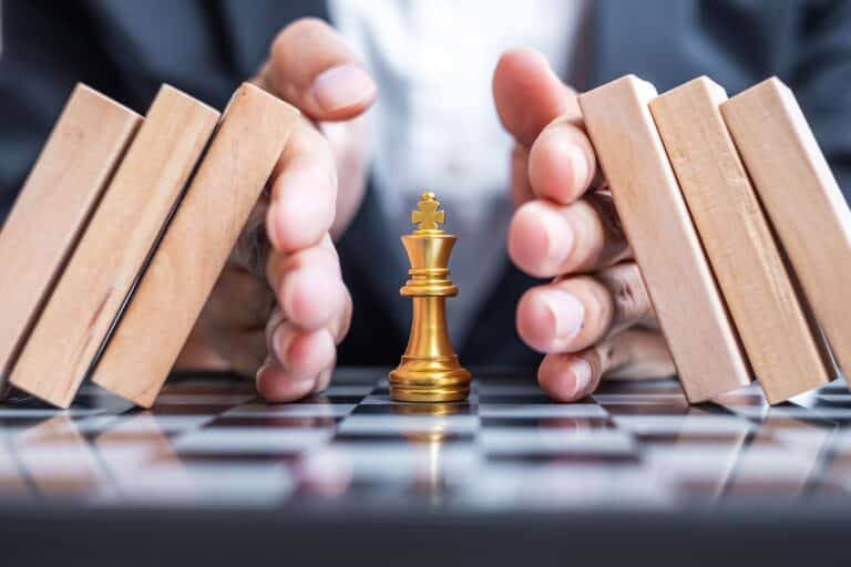 Hands shielding a chess piece as falling dominos approach, symbolizing protecting value and avoiding business transaction pitfalls